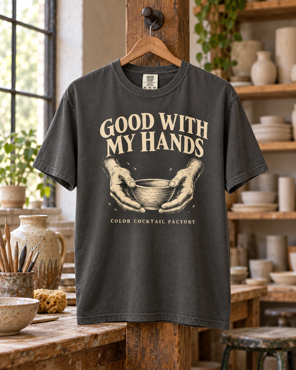 Pepper charcoal Good With My Hands pottery T-shirt with cream hands-and-bowl artwork, hanging in a sunlit pottery studio.